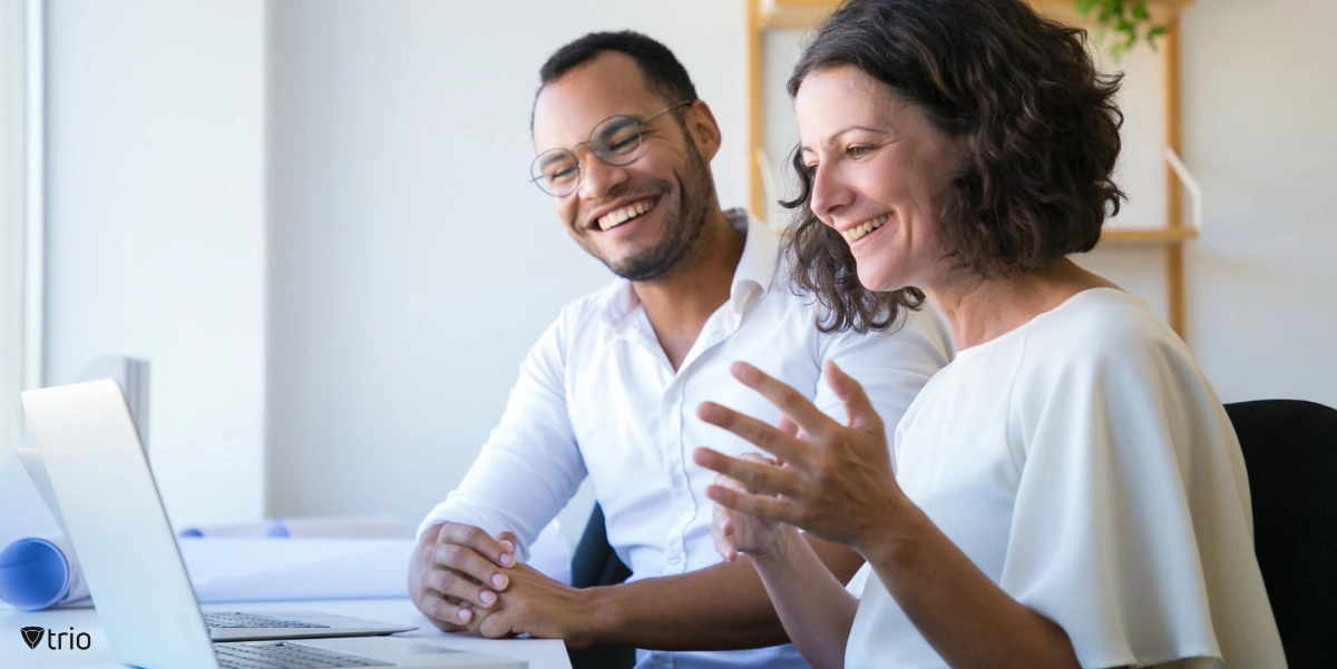 A man and a woman, both smiling and engaged in a cheerful conversation at a bright office desk with laptops. 