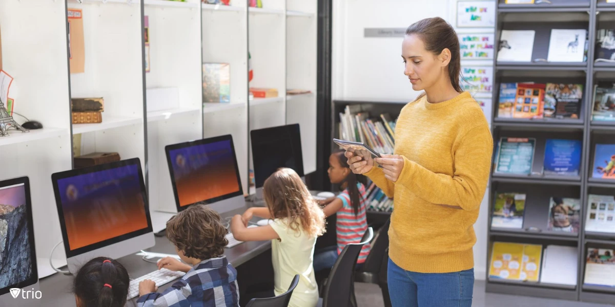 A teacher stands in front of a row of young students using desktop computers, holding a tablet to monitor their activities, in a school library or classroom, with bookshelves in the background