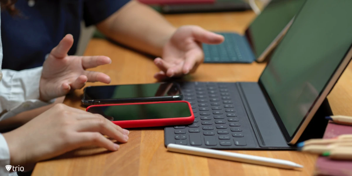 Two people’s hands rest on a wooden table, near a tablet with a keyboard attachment and multiple smartphones