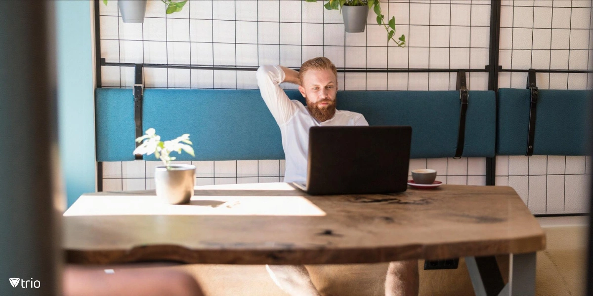 A bearded man with light-colored hair sitting at a wooden table, wearing a white shirt and working on a laptop while leaning back with one hand resting behind his head