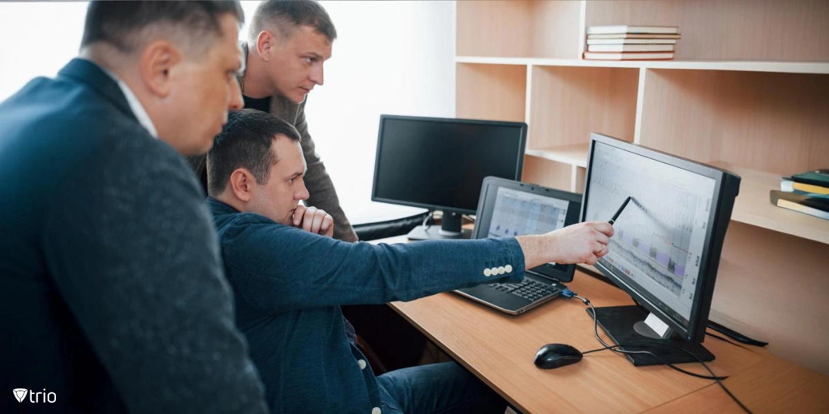 Three people are in an office setting working together. One person points to a computer monitor, appearing to explain or analyze data displayed on the screen