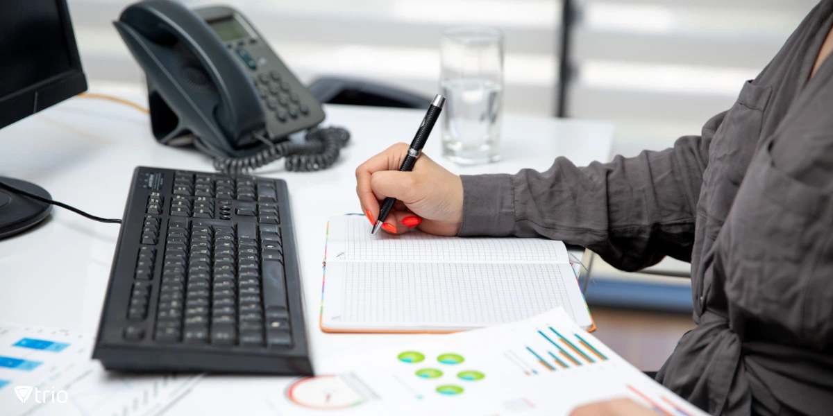A business professional seated at a desk, taking notes in a notebook while analyzing printed graphs and charts.