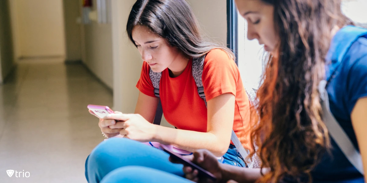 Two teenage girls, dressed casually with backpacks, sit on the school hallway floor while focused on their smartphones