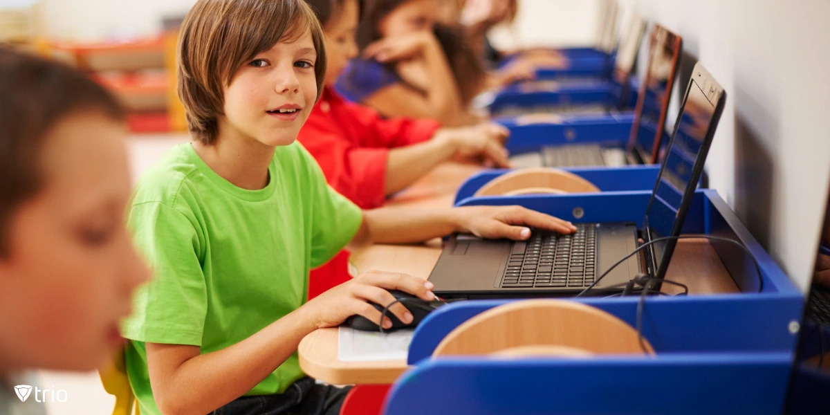 A boy in a bright green shirt is smiling while using a laptop in a classroom with other students. The students are seated at wooden desks with blue dividers, each equipped with a laptop. The background shows more children engaged in their work