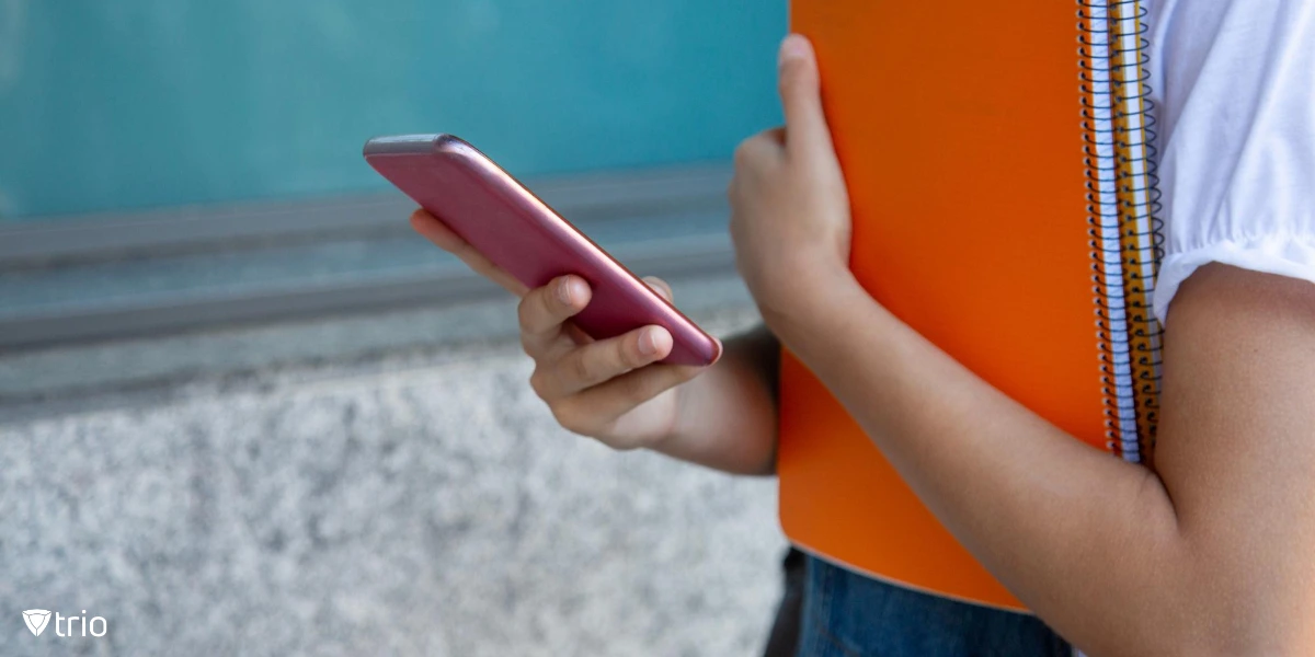 A student in casual attire holds an orange notebook while using a pink smartphone, seemingly engaged in a digital activity outside a school building.