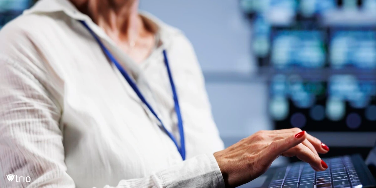 A professional woman in a high-tech facility, wearing a lanyard, types on a laptop, possibly managing IT systems or network security.