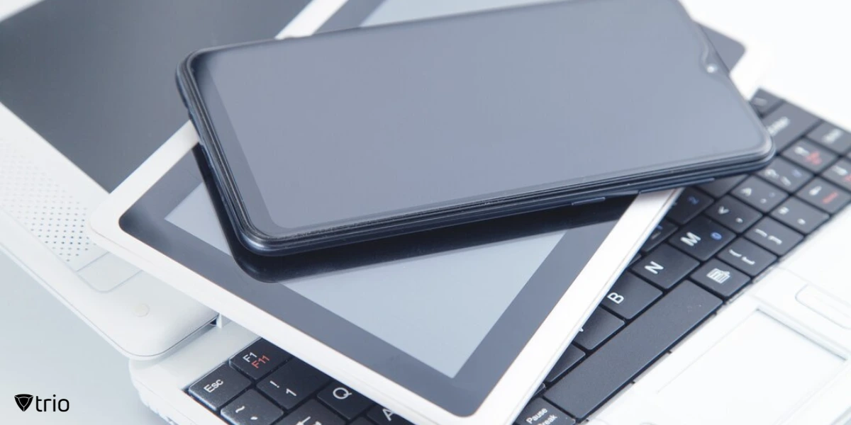 A modern flat lay of multiple smart devices stacked on top of each other. A smartphone is placed over two tablets, which are resting on an open laptop keyboard.