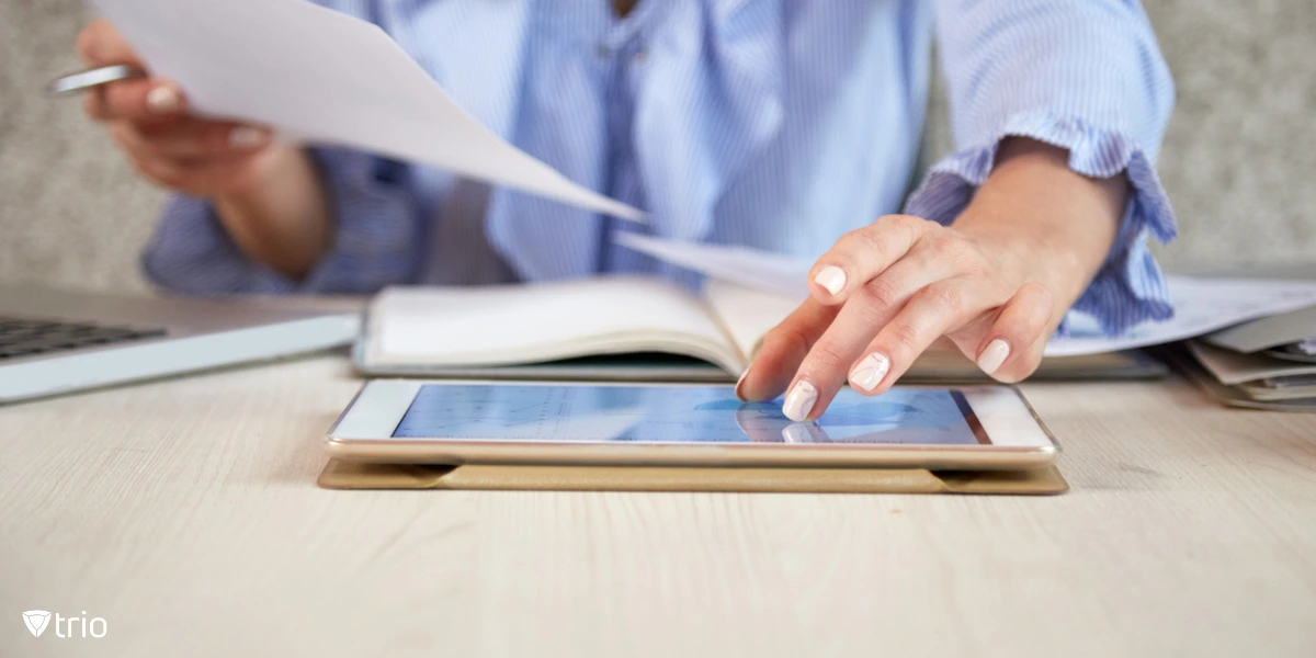 [A woman in a blue blouse is multitasking at a desk, using a tablet while handling paperwork.