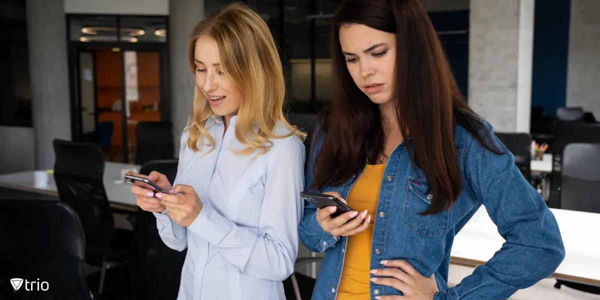 Two coworkers distracted by social media on their smartphones in an office environment.